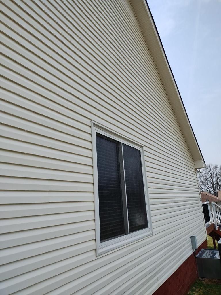 Side view of a beige house with vinyl siding and a double window under a cloudy sky.