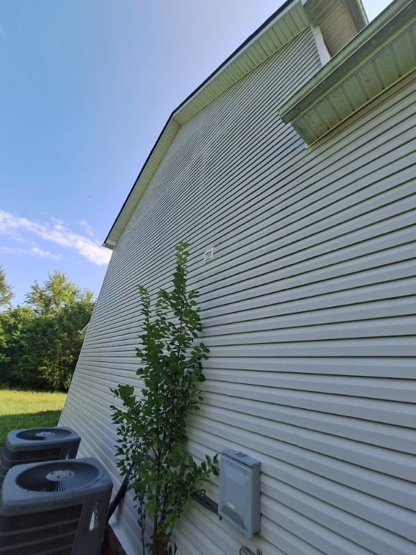 Side of a white house with vinyl siding, shrubs, and two air conditioning units by a grassy yard.