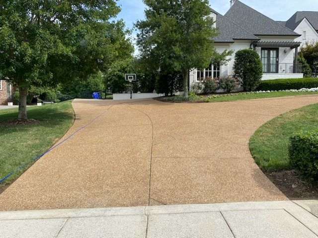 Curved tan driveway leading to a gray house with trees and trimmed shrubs