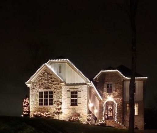 House outlined with white Christmas lights at night, with illuminated trees and a dark sky.