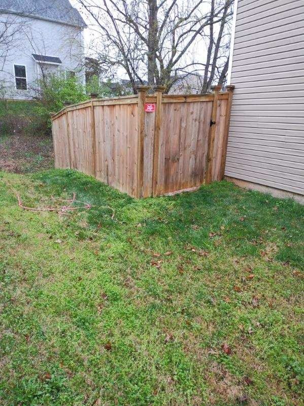 Wooden corner fence in a grassy backyard beside a beige house, with leafless trees beyond.