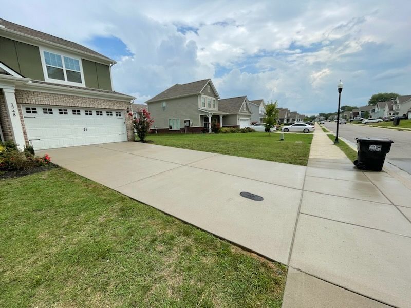 Suburban driveway and sidewalk beside a house, with a grassy lawn and trash bin on the curb.