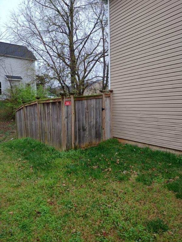 Backyard corner with weathered wooden fence, beige house siding, grass, and leafless trees
