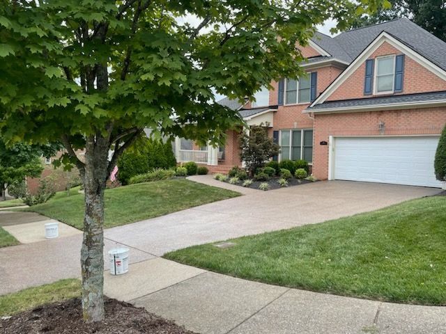Suburban brick house with a two-car garage, driveway, and front lawn viewed from the sidewalk.
