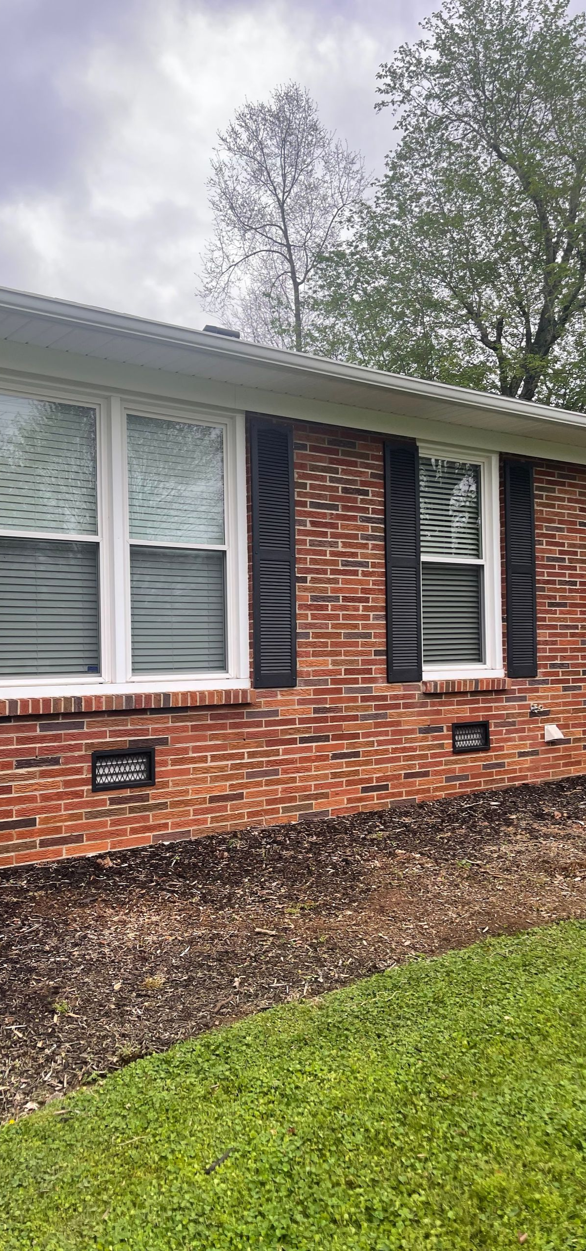 Brick house exterior with white windows, black shutters, and green lawn under overcast sky