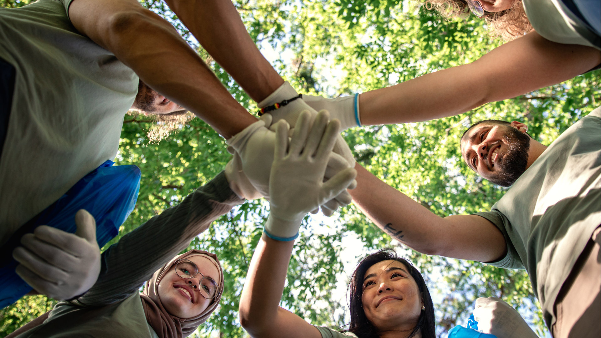 A group of diverse volunteers wearing gloves stack their hands in a circle against a background of green trees.