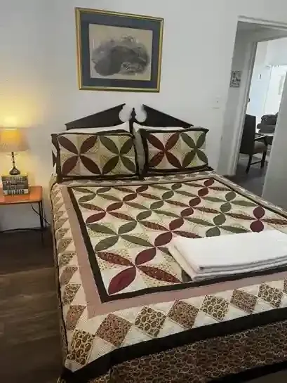 A bed with a quilt featuring a geometric floral pattern in red, green, and tan, with a folded white towel on top.