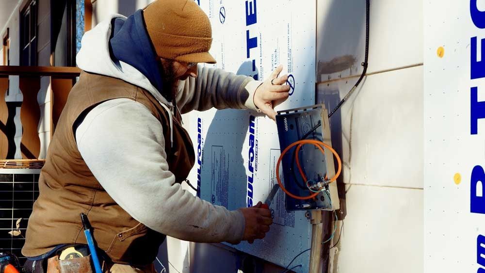 Man in brown vest working on exterior wall, cutting around an electrical box.