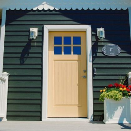 Tan door with square window, surrounded by white trim, on dark green siding, white scalloped roof, with a flowerpot.