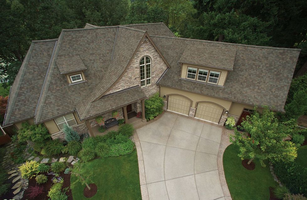 Aerial view of a large, tan house with stone accents, a driveway, and lush landscaping surrounded by trees.