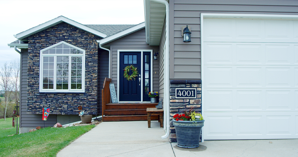 House exterior with gray siding, stone accents, and white garage door; a front door with a wreath is visible.