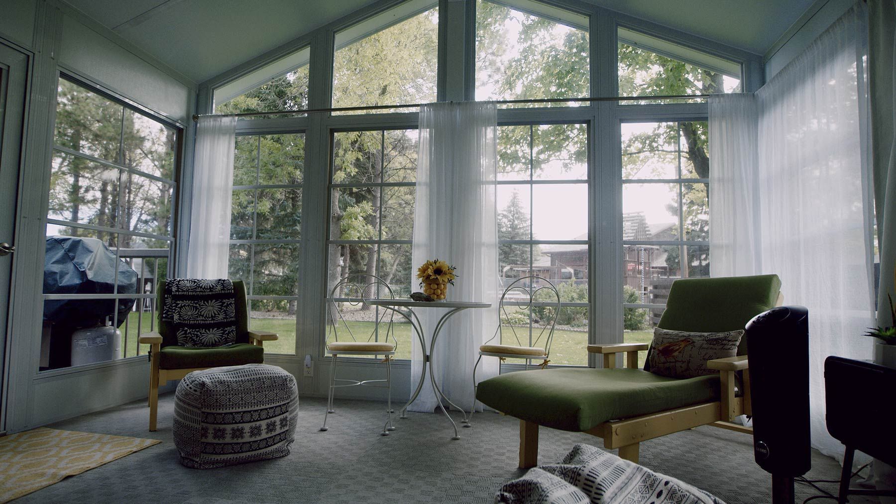 Sunroom with green chairs, small table, and large windows overlooking a yard.
