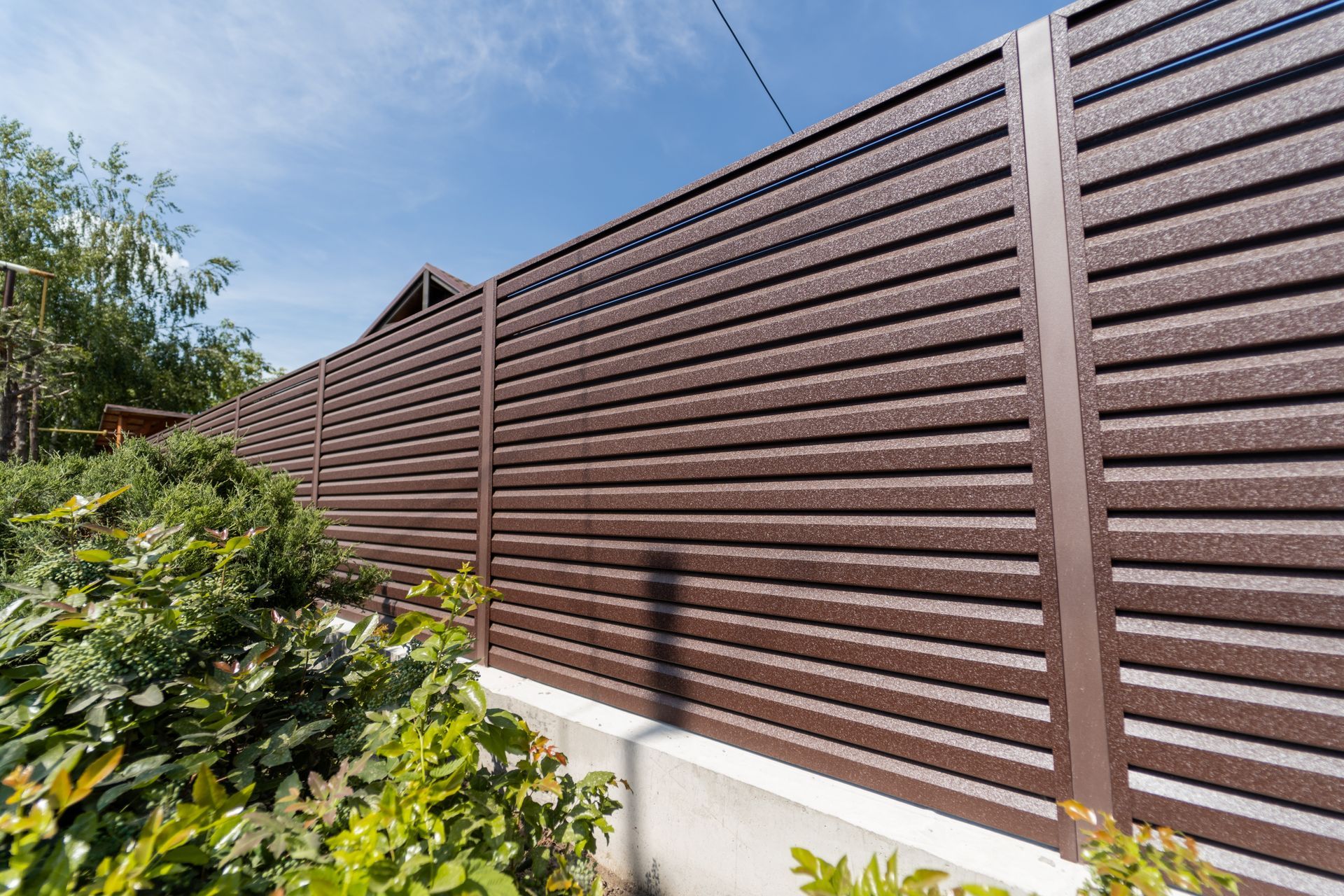Brown horizontal slatted fence with vertical supports in a sunny outdoor setting, with plants and blue sky visible.