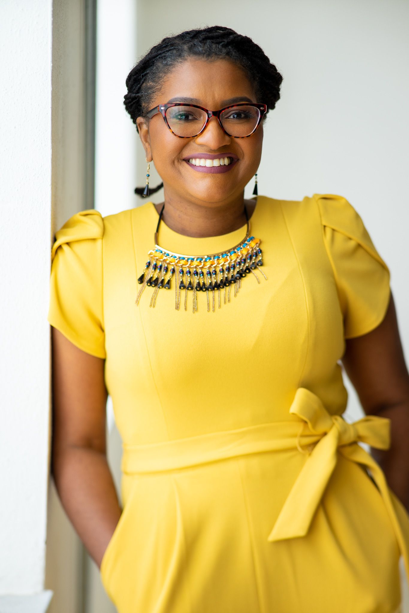Woman in yellow dress, smiling, wearing glasses and necklace, hands in pockets. Leaning against white wall.