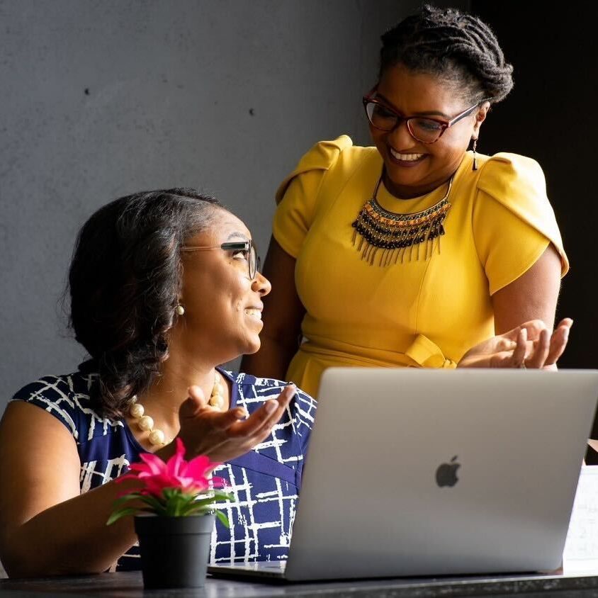 Two women at a laptop, one points, smiling. One in blue top, other in yellow dress. Dark interior, desk.