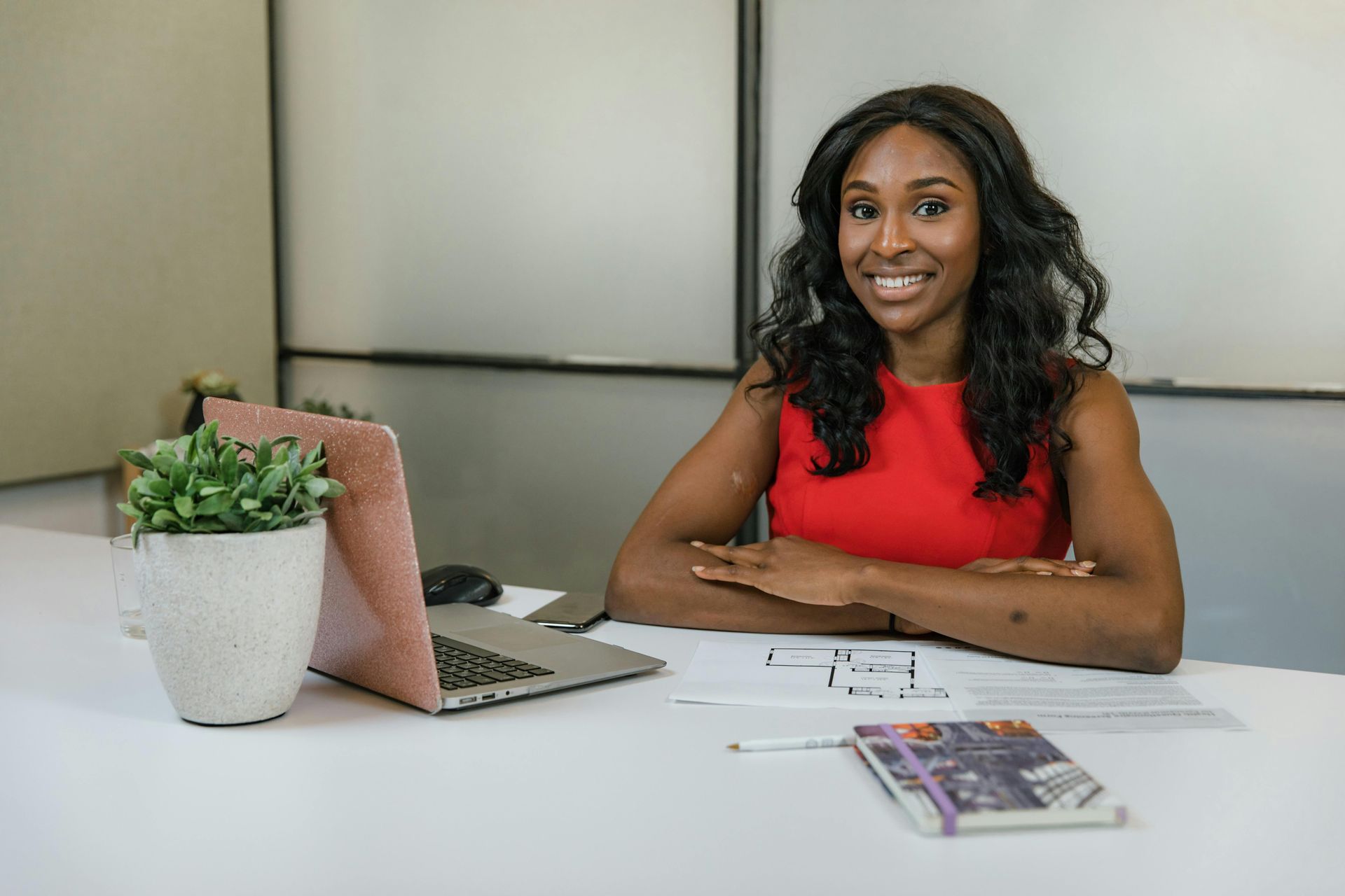 Woman with dark hair, red dress, smiling, seated at a desk with laptop, notebook, and plant.