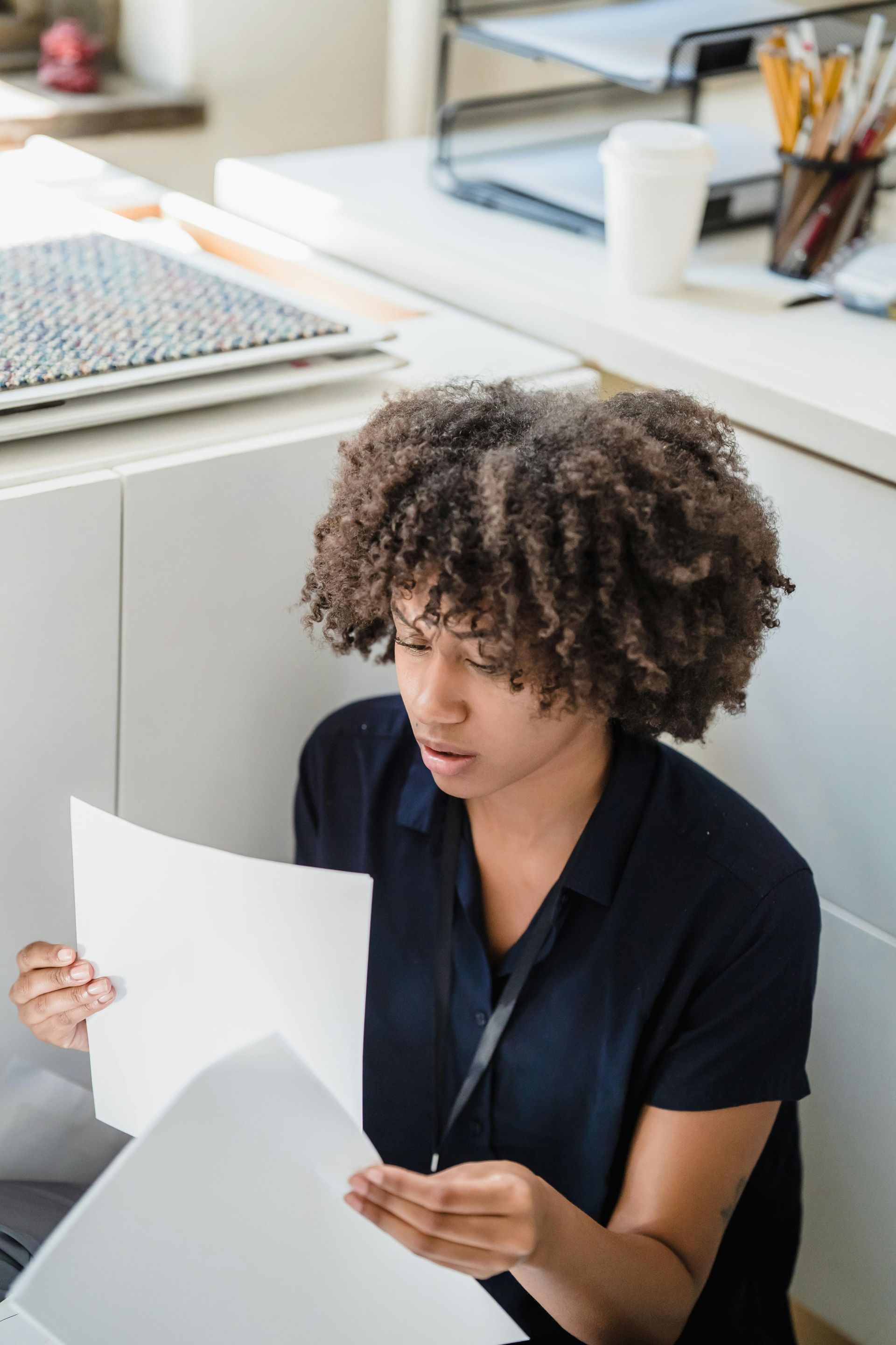 Woman with curly hair examines documents, seated in a white office space, looking concerned.