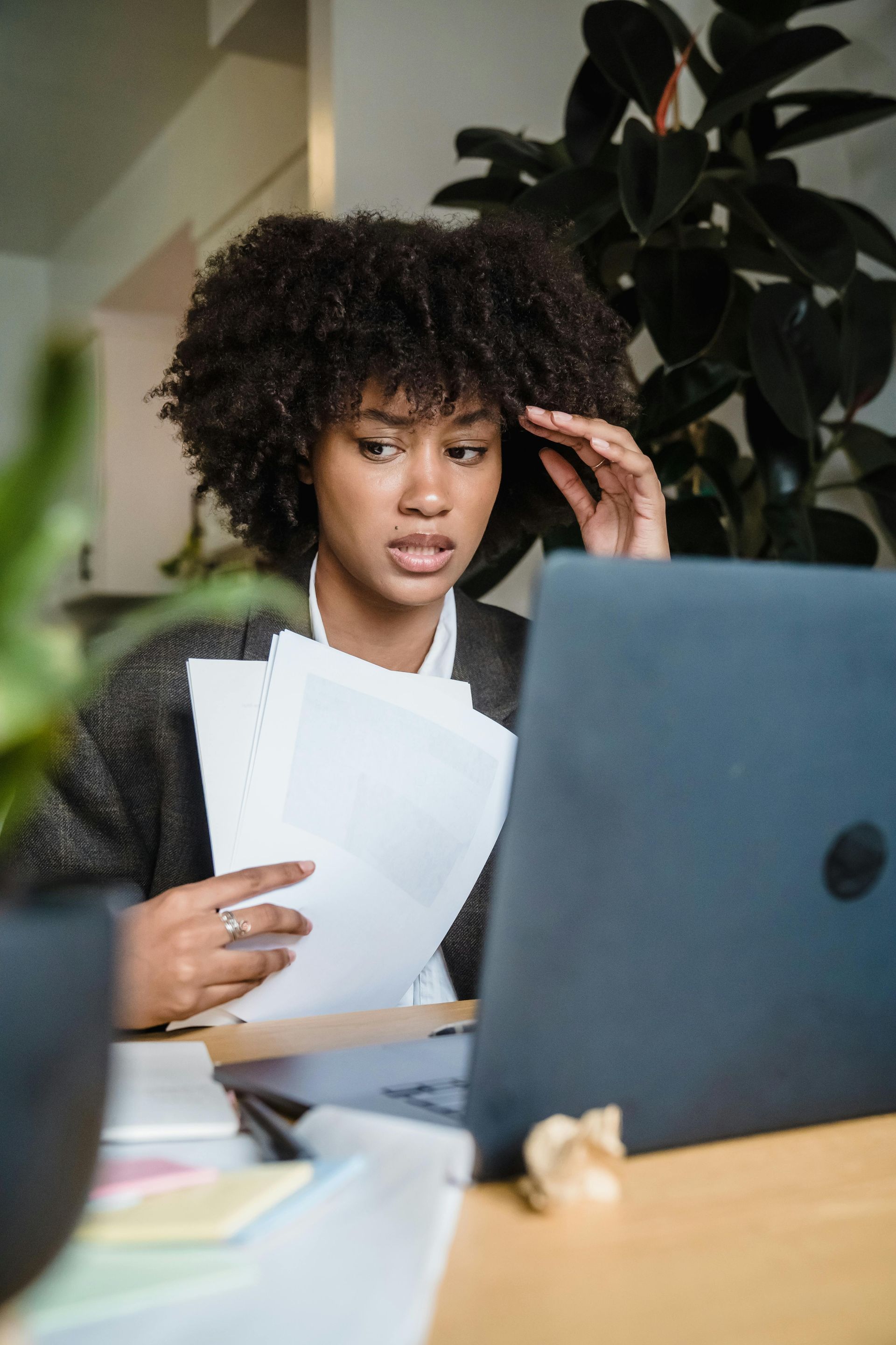 Woman with dark curly hair, stressed, looking at laptop, holding papers, home office setting.