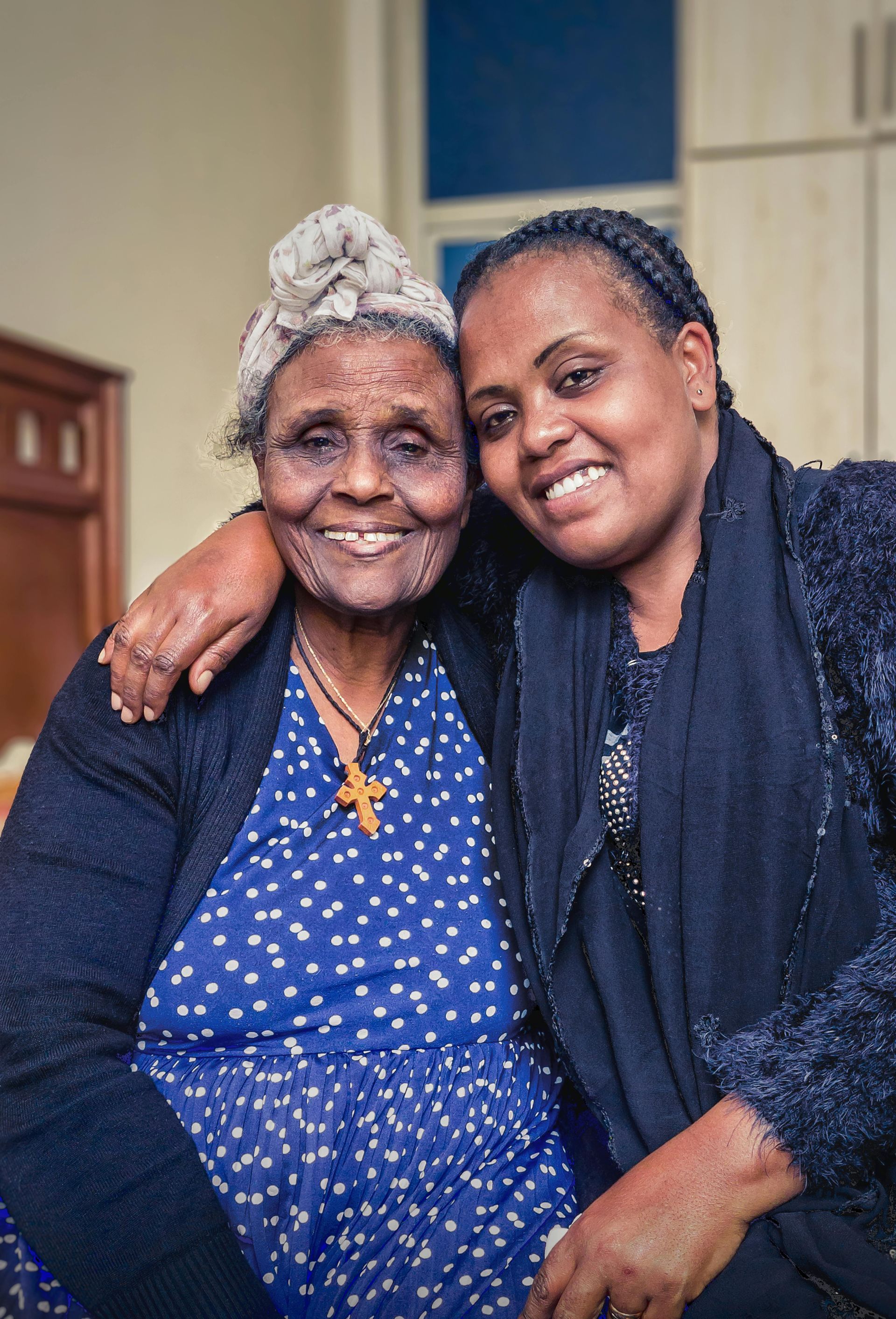 Older woman in blue dress and younger woman in black embracing, both smiling indoors.