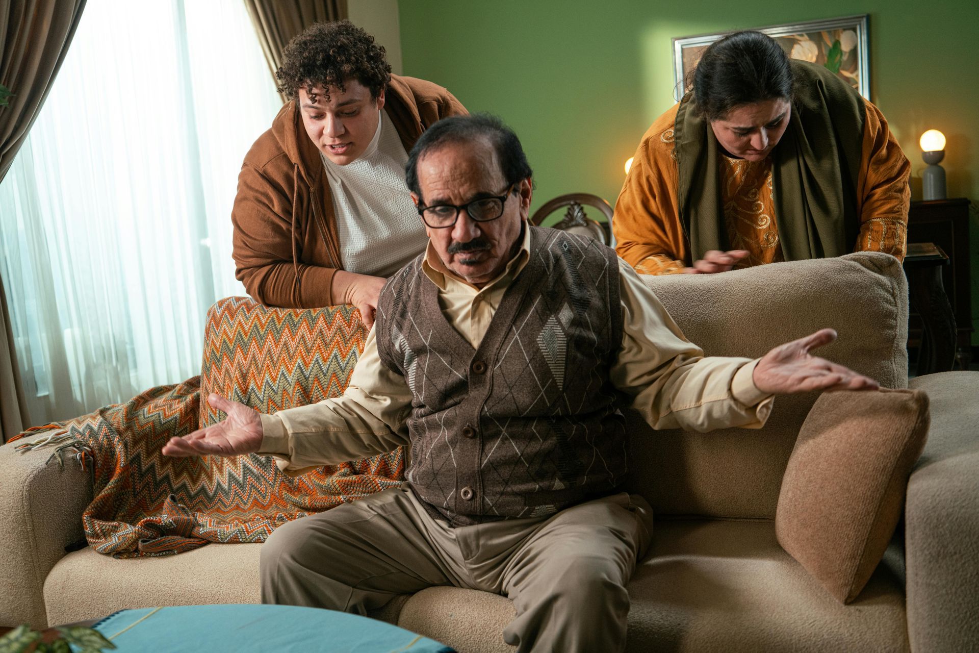 Man seated with hands out, looking perplexed. Two people lean over the couch. Green wall, neutral-toned couch.