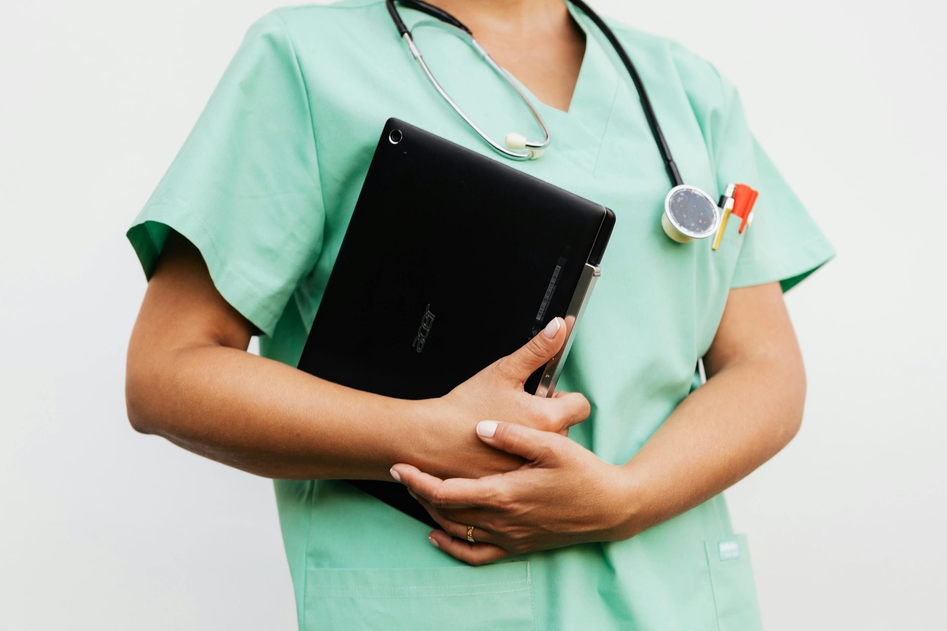 A healthcare professional in mint-green scrubs holds a black tablet and wears a stethoscope around their neck.