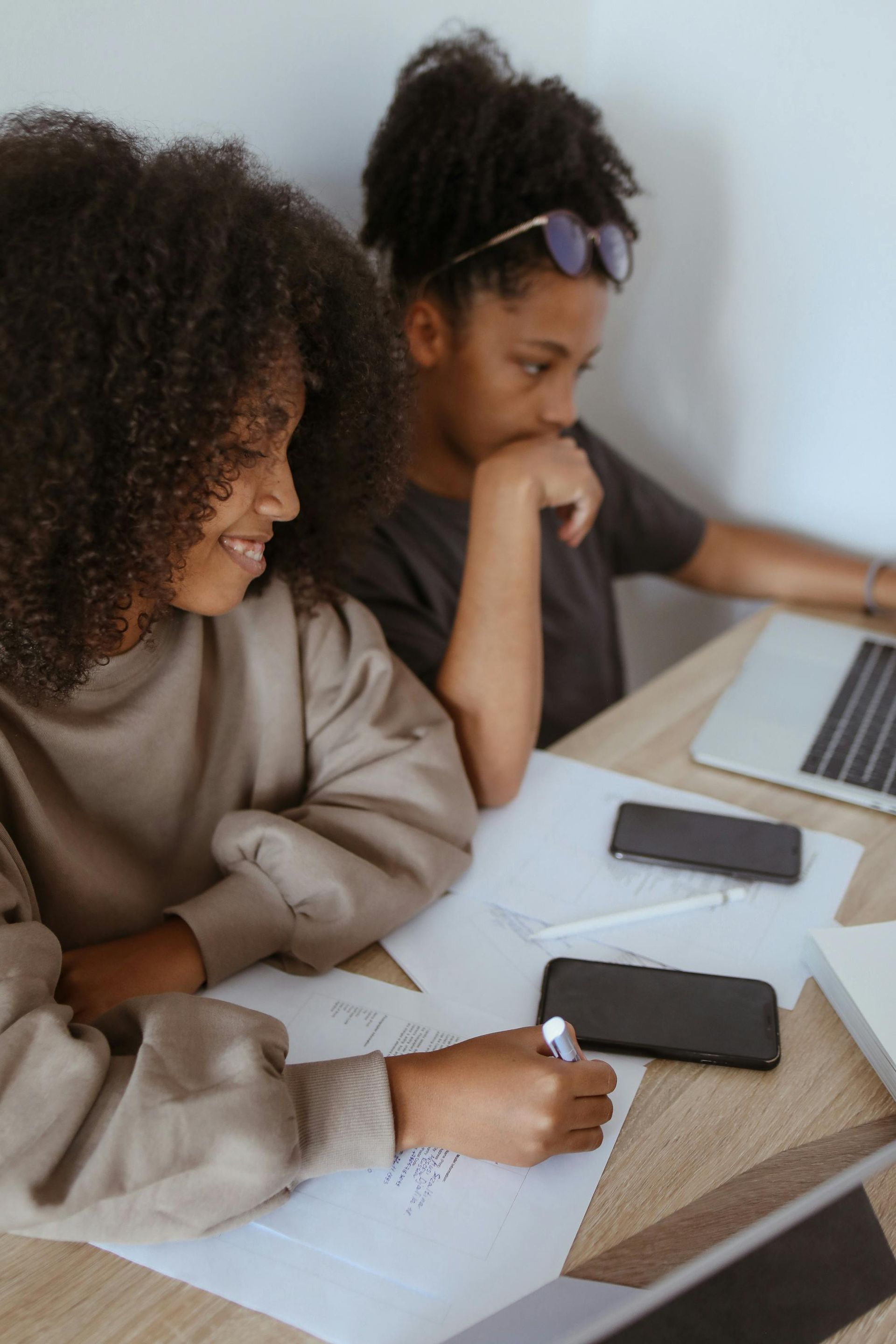 Two students work together at a desk, one writing, the other using a laptop.