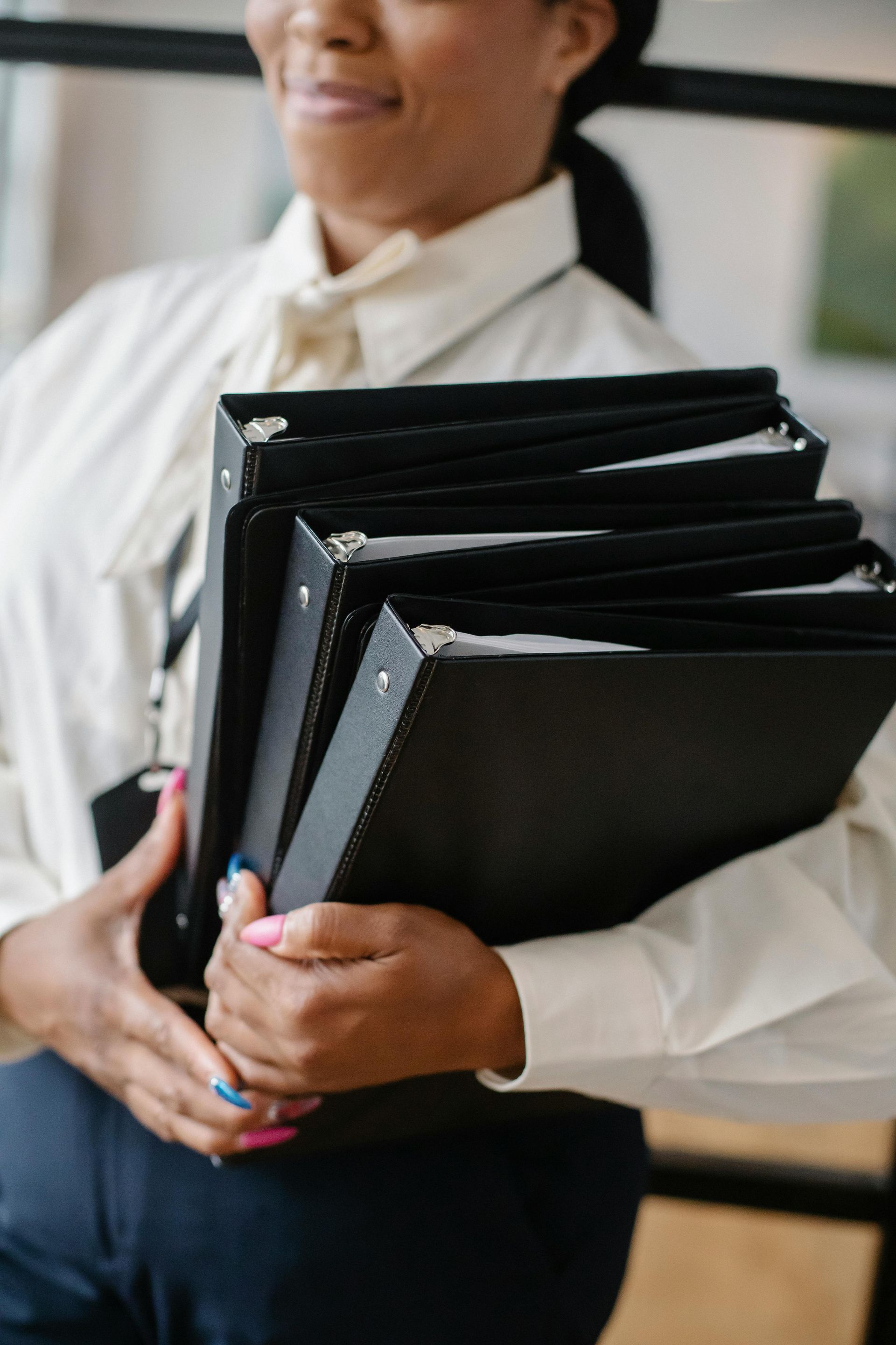 Person in a white shirt and navy pants holding several black binders.