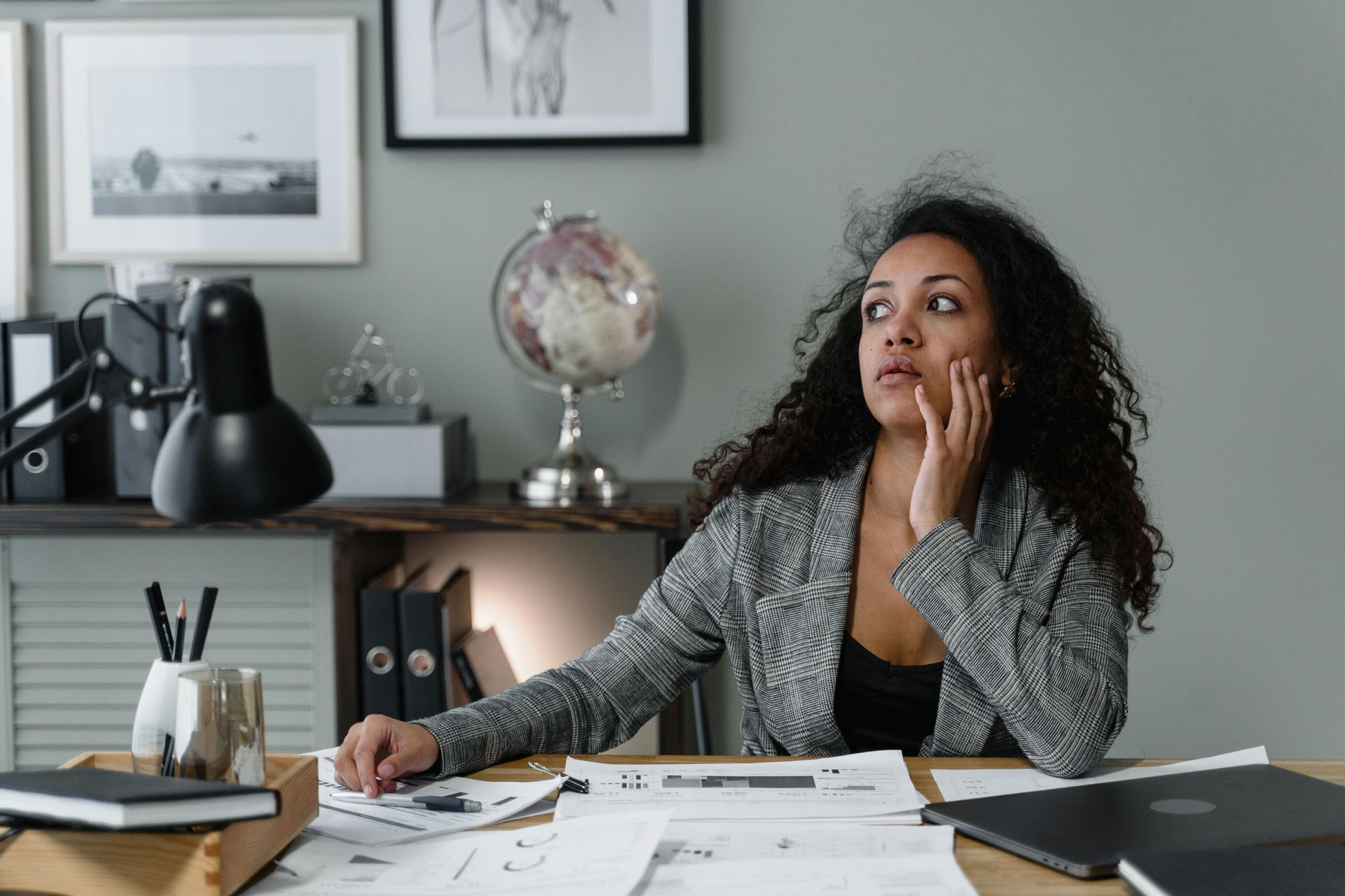 Woman with curly hair looks upward, resting her head on her hand while sitting at a desk with papers and a globe.