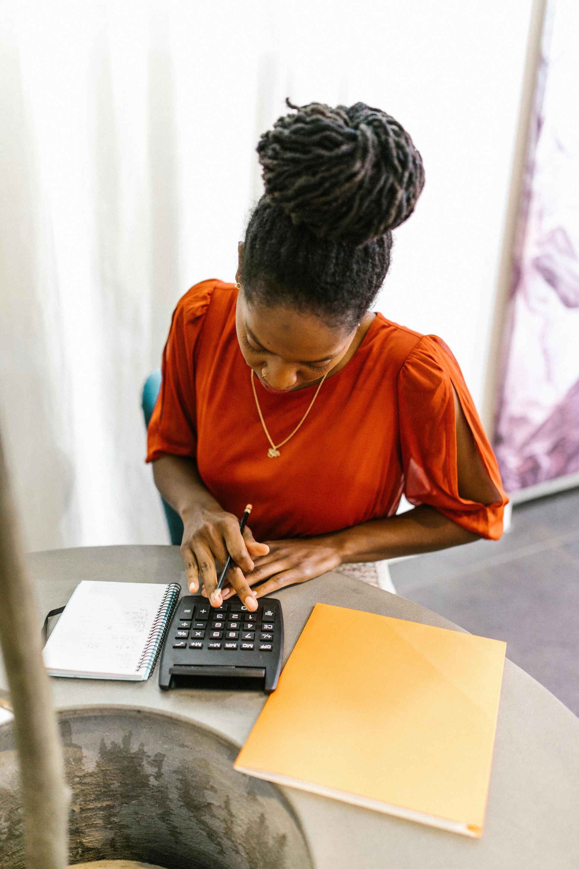 Woman with dark hair bun wearing red shirt, using a calculator at a table with notepad and file.
