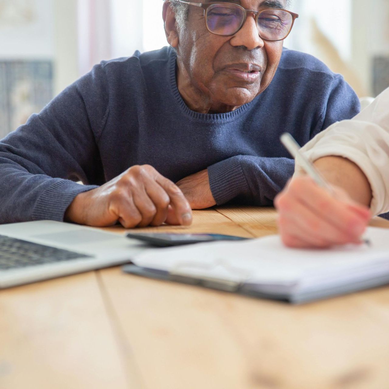 Senior man and assistant reviewing documents, taking notes at a wooden table with laptop.