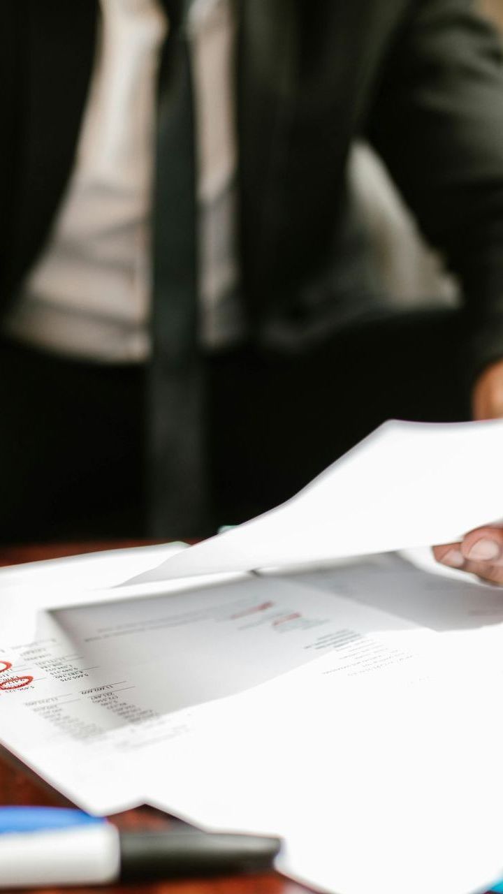 Person in suit handing paperwork to another person, documents on desk.