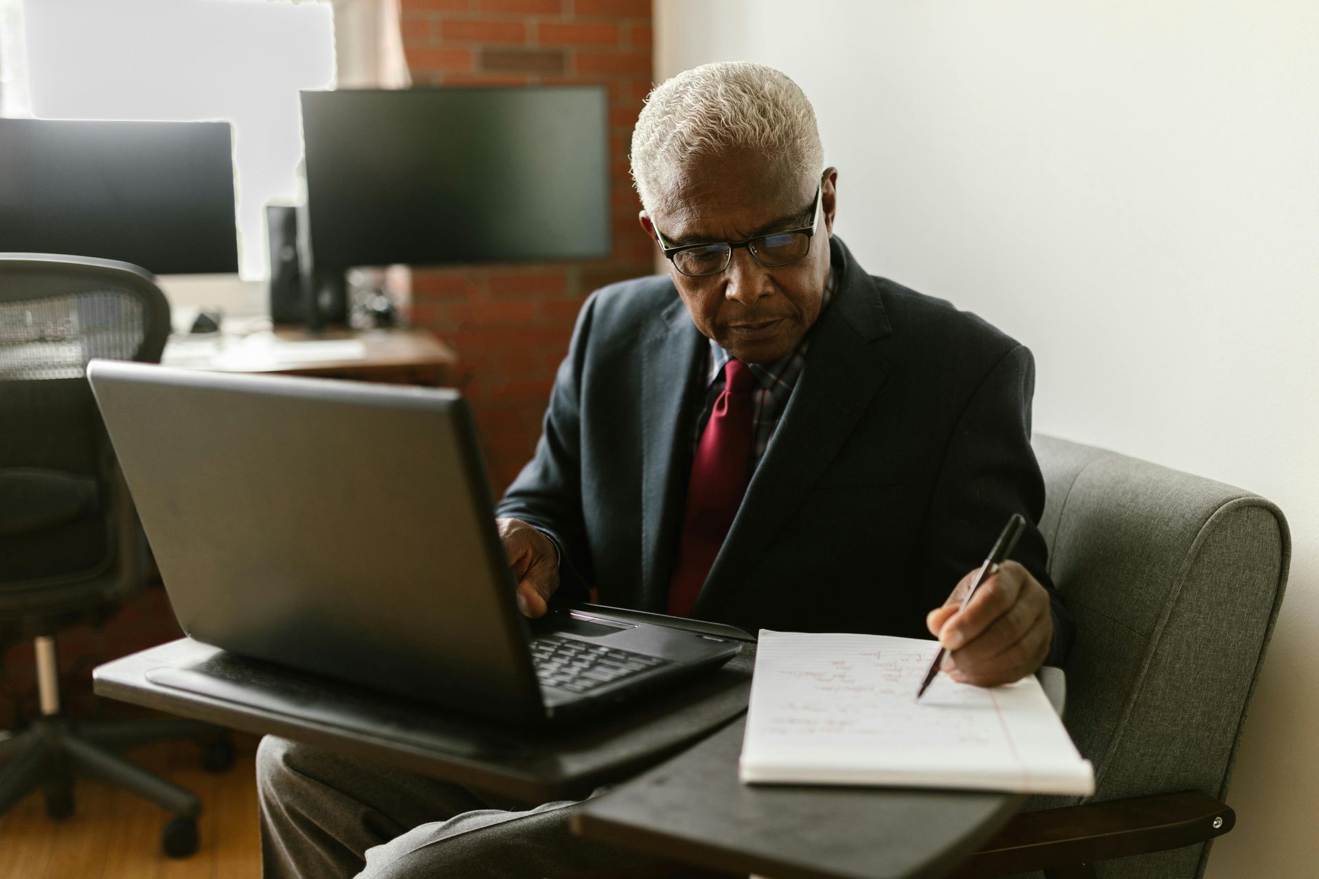 An older Black man in a suit, writing on a pad while using a laptop, indoors.