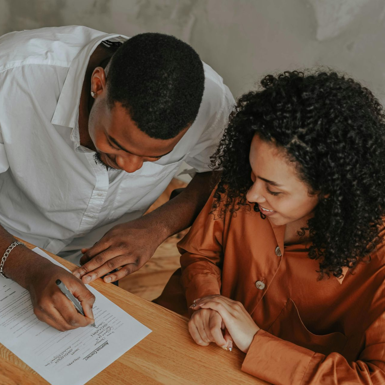 Couple signing document at a table, man writing, woman smiling, indoors.