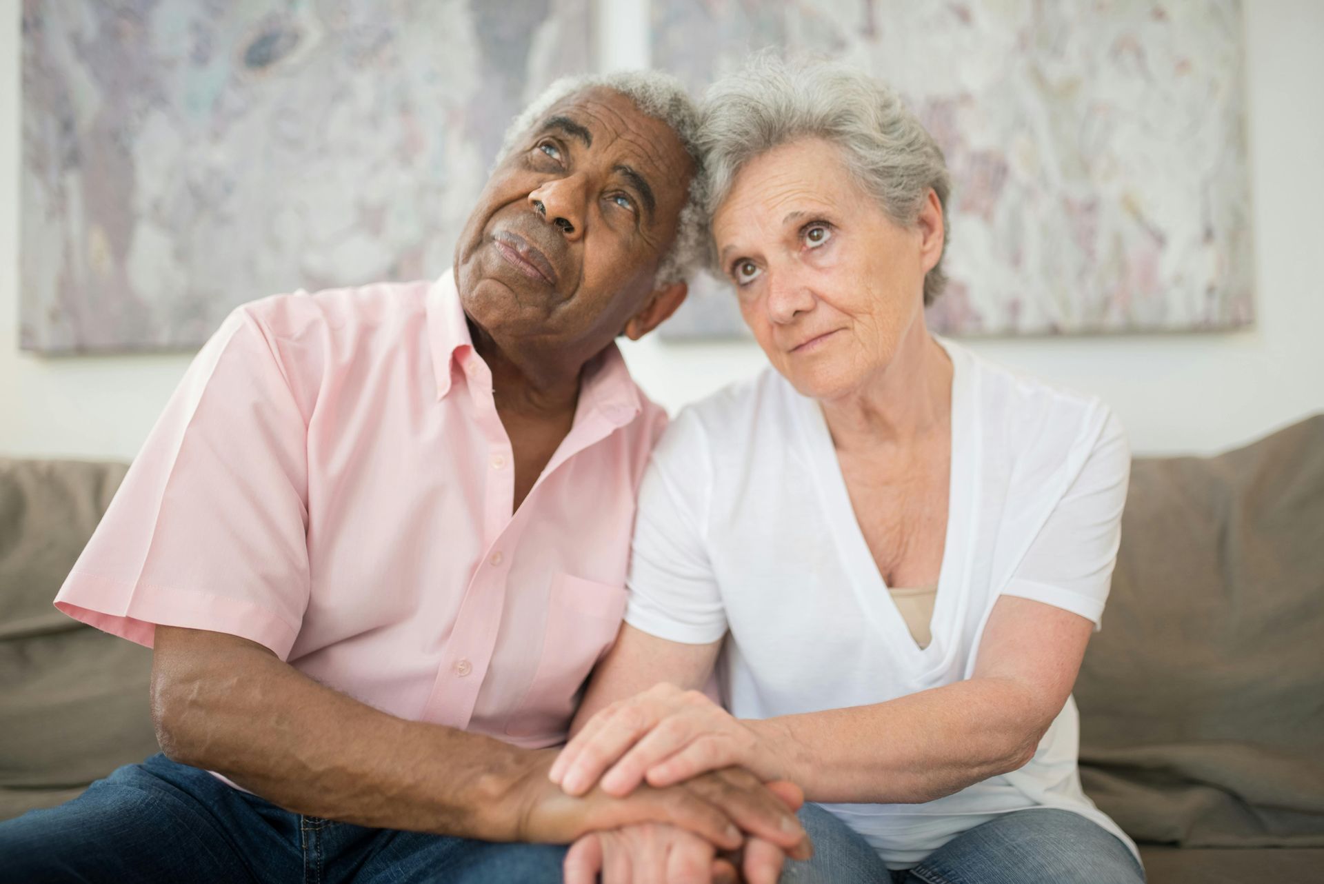 An elderly couple sits close together, holding hands. Both are looking upward with pensive expressions.