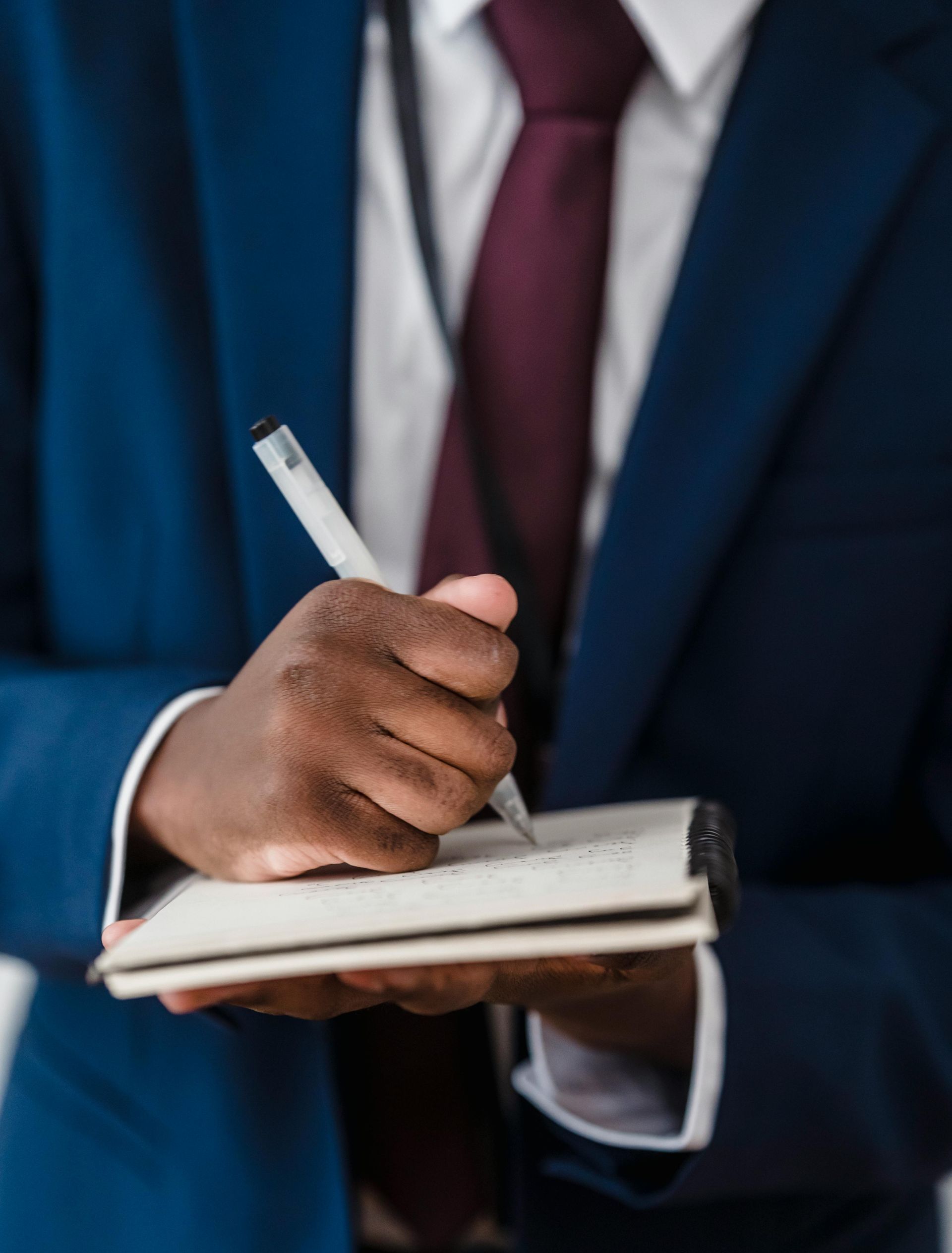 Black man in blue suit writing in notebook, wearing a red tie.