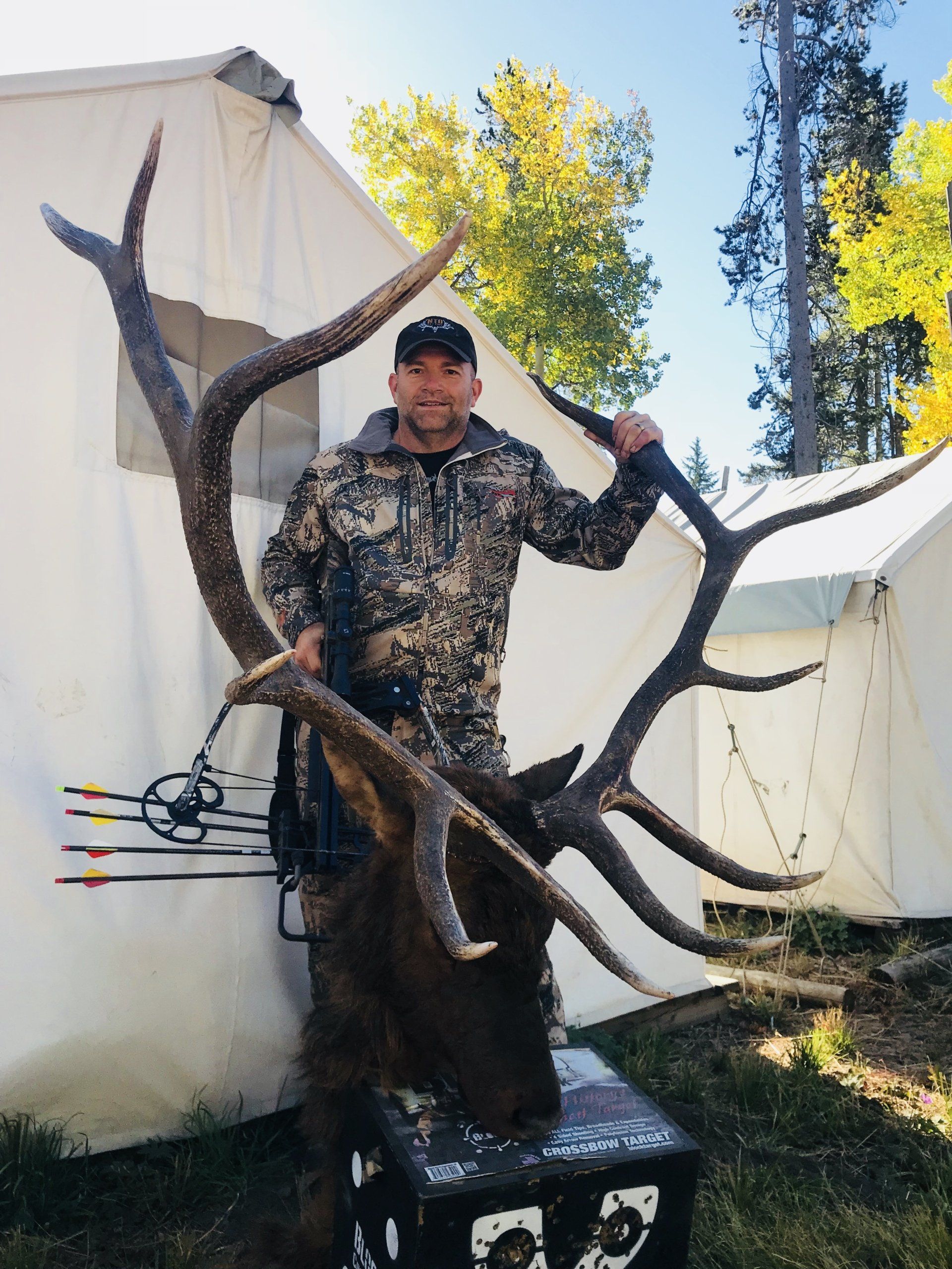 Man in camo holds large elk antlers, posing next to a tent.