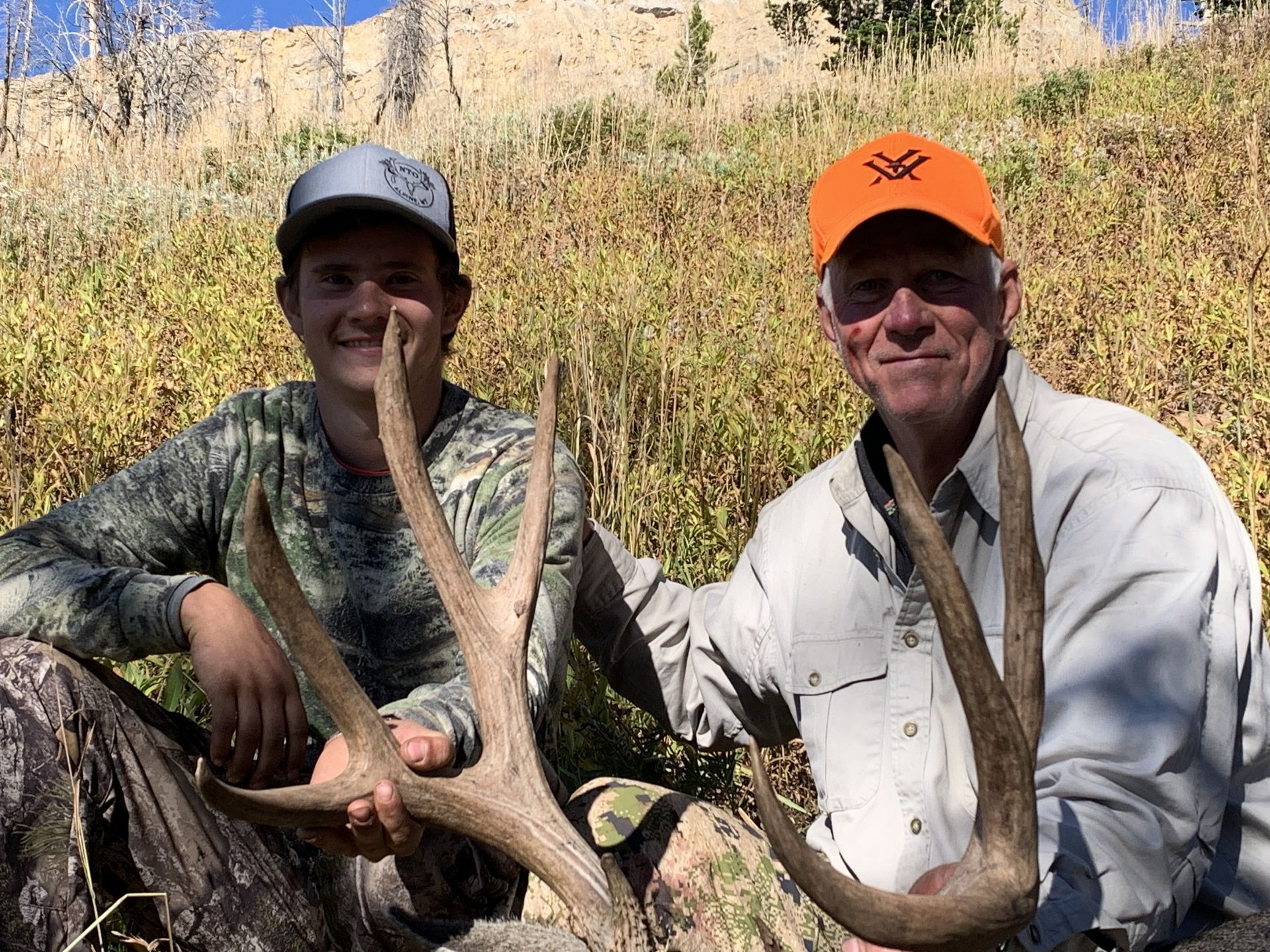 Two people with a deer's large antlers pose outdoors. One wears a camo shirt, the other an orange hat.
