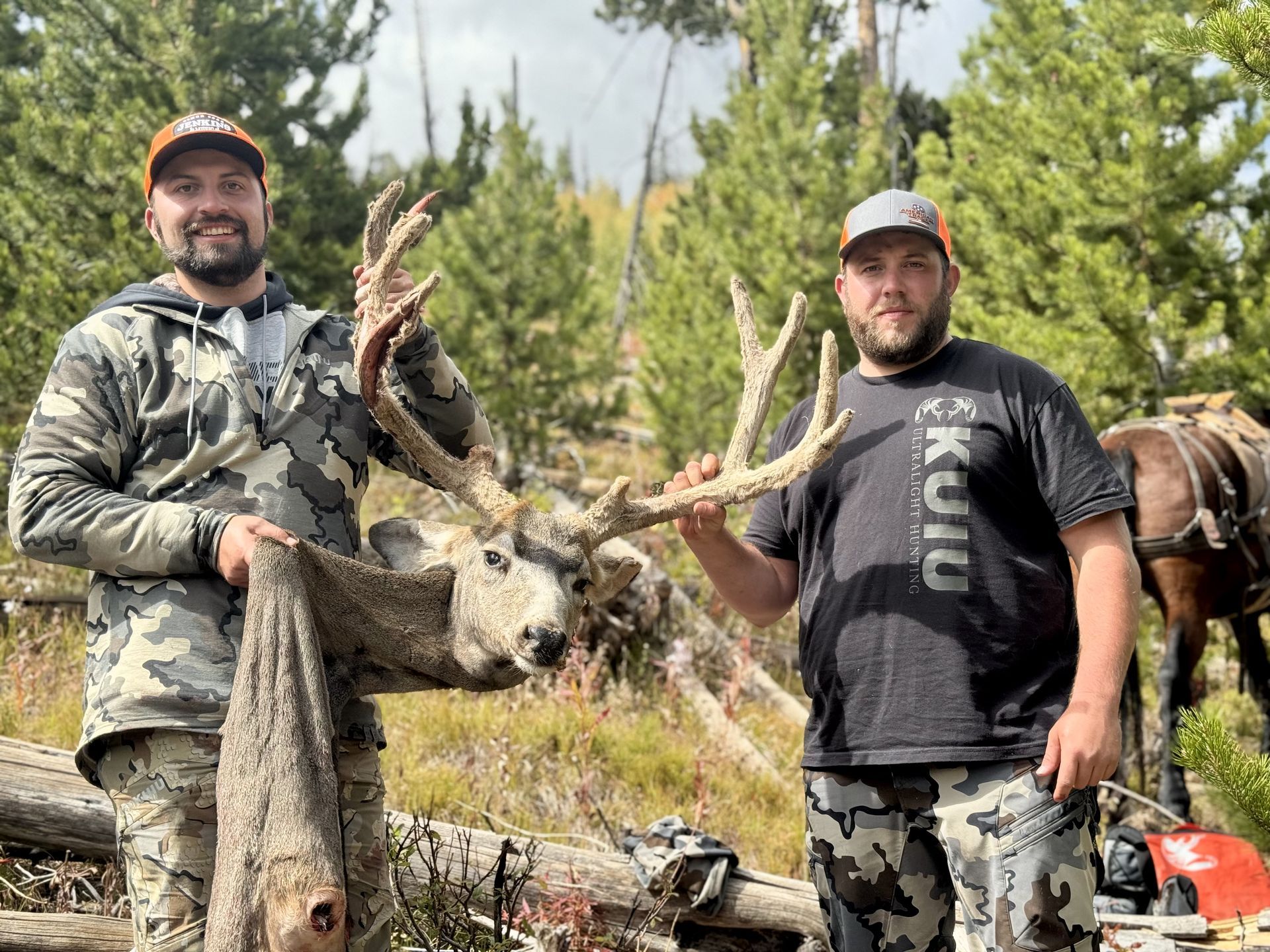 Two men hold a large deer with antlers in a wooded area. They wear hunting attire and smiles.