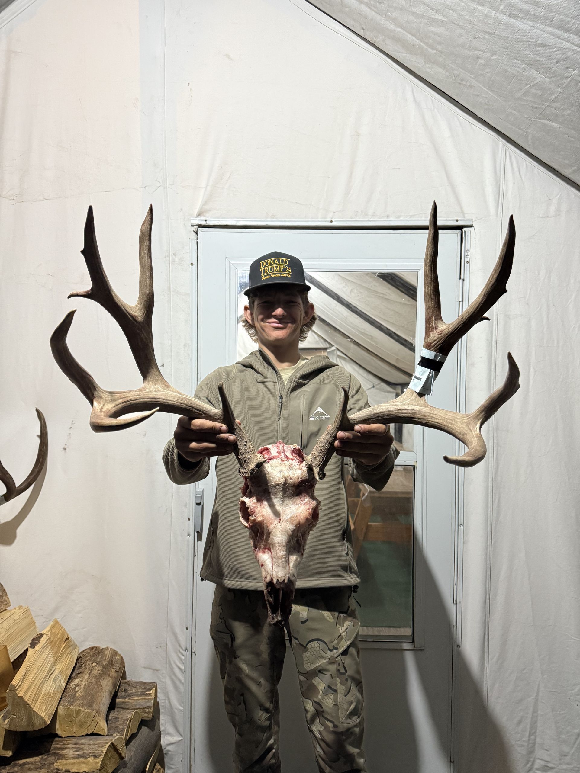Man holding large deer antlers, indoors near a doorway and firewood, wearing a hat and camouflage.