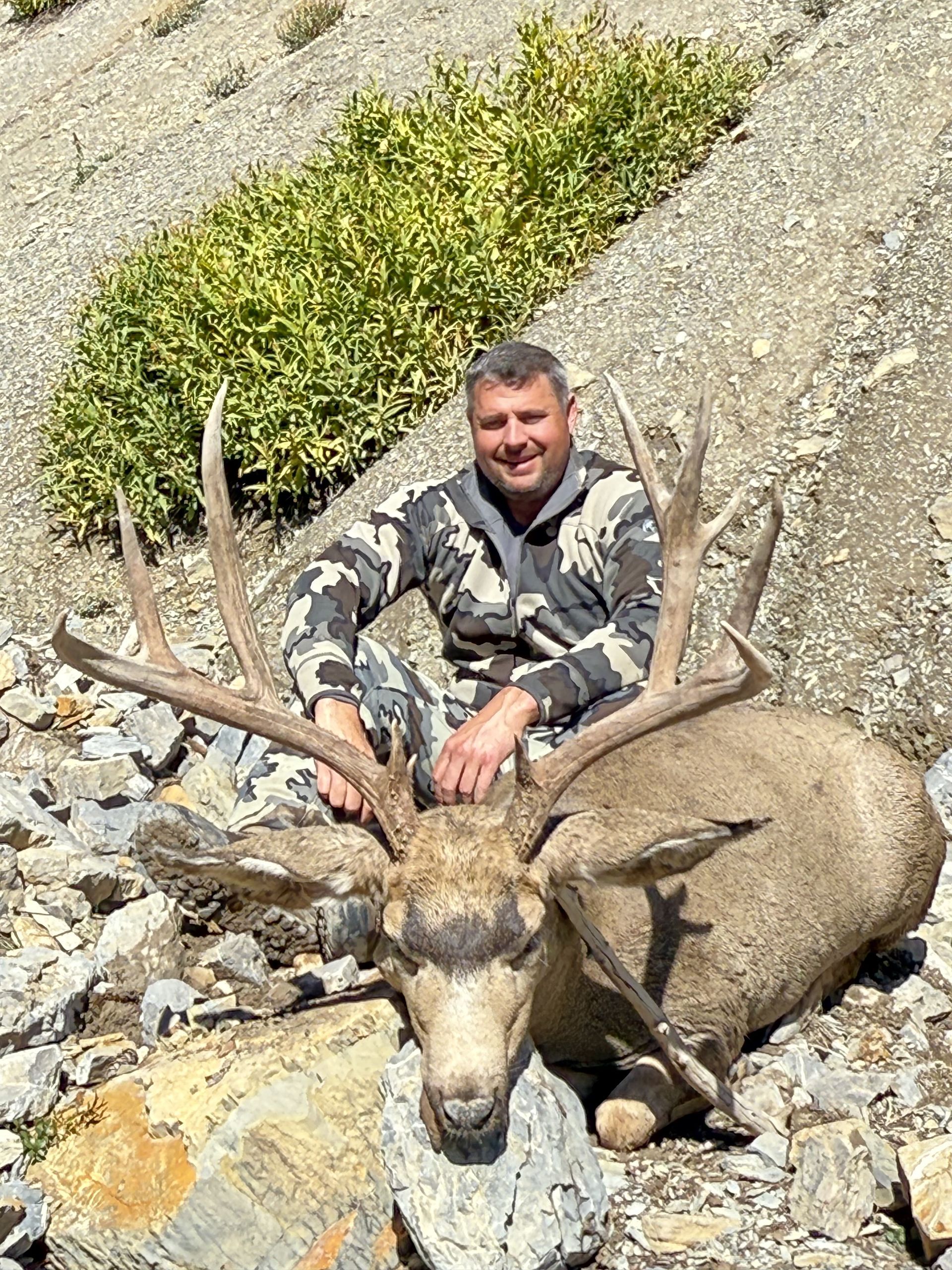Man in camo with large buck, Rocky Mountain setting.