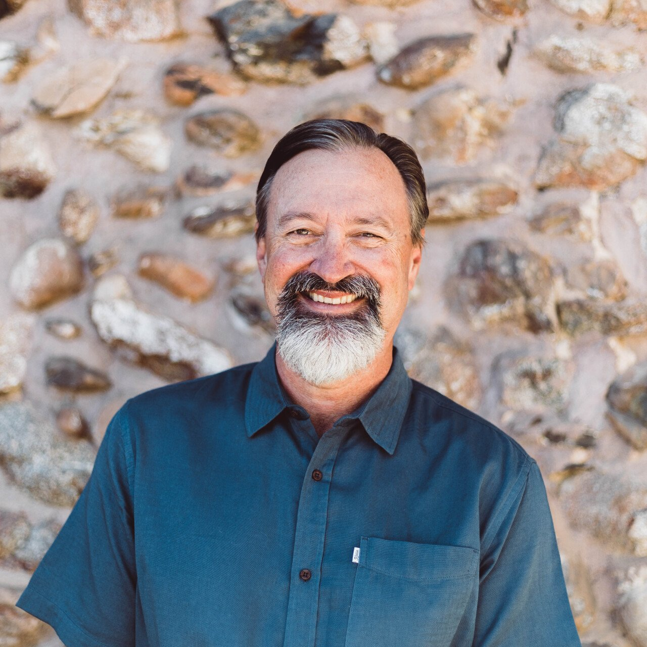 A man with a beard is smiling in front of a stone wall