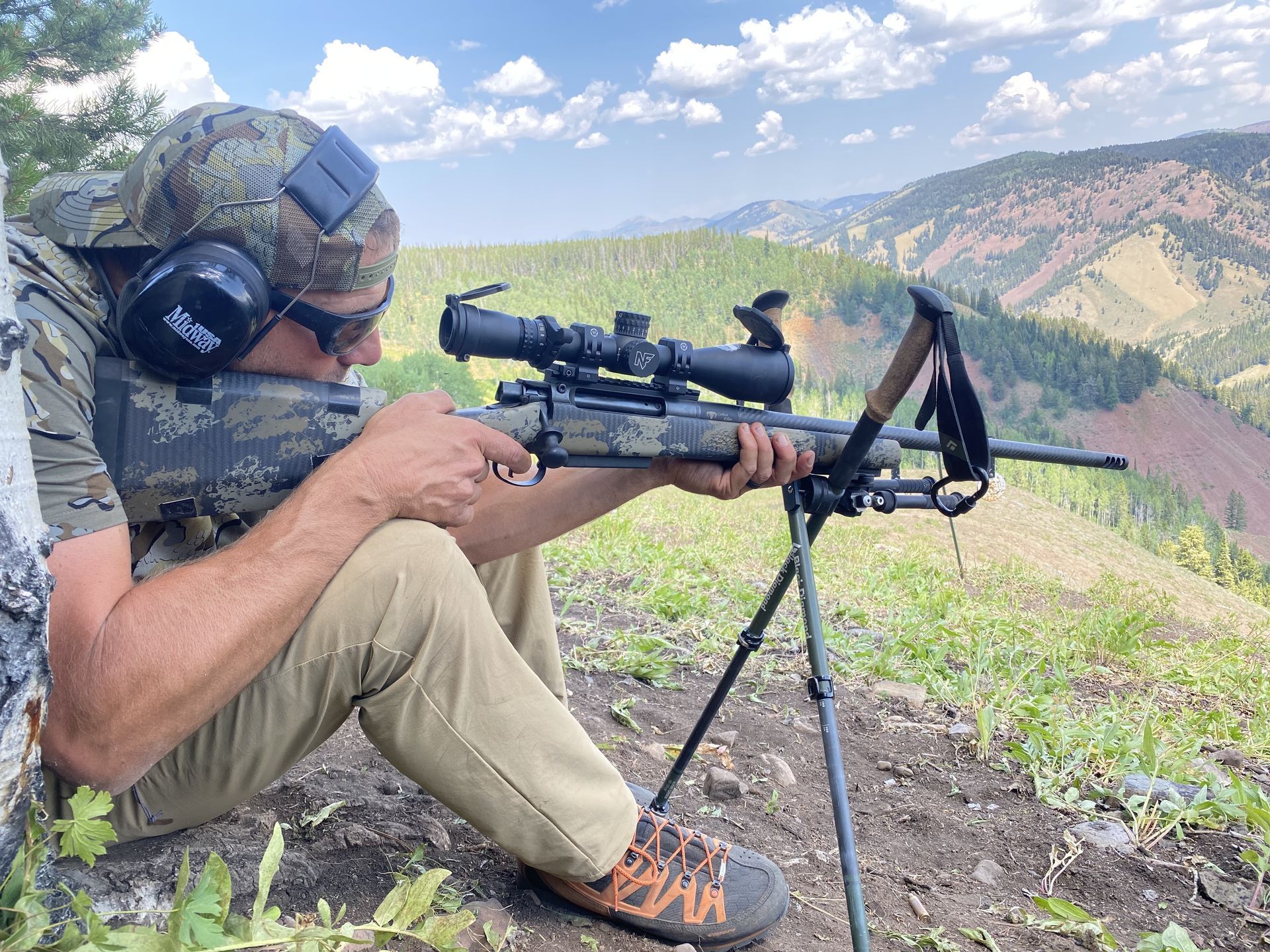 Person aiming rifle on a tripod in a mountainous setting, wearing camouflage, ear protection, and sunglasses.