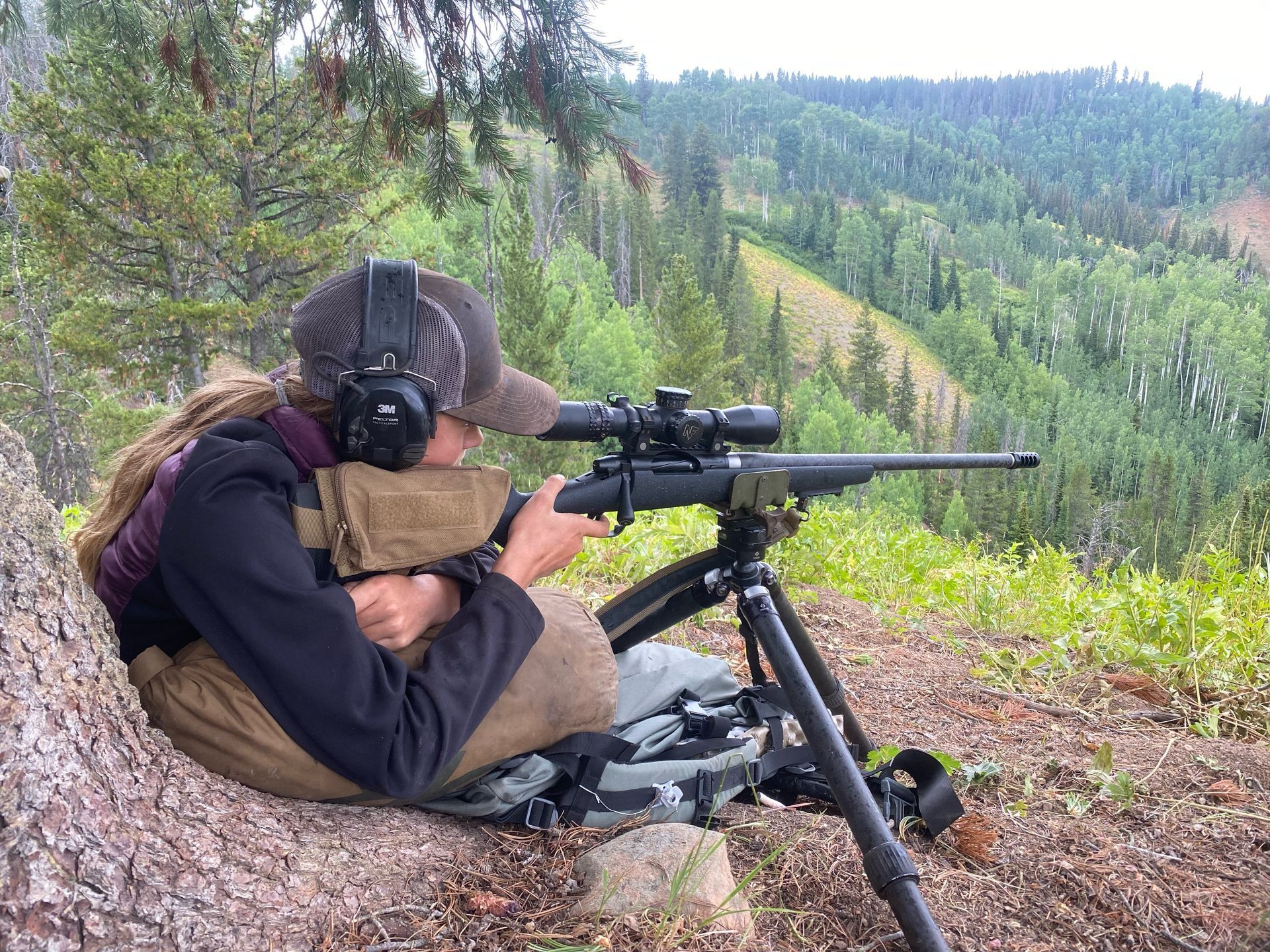Person aiming a rifle with scope, using a tripod, outdoors in a forested area.