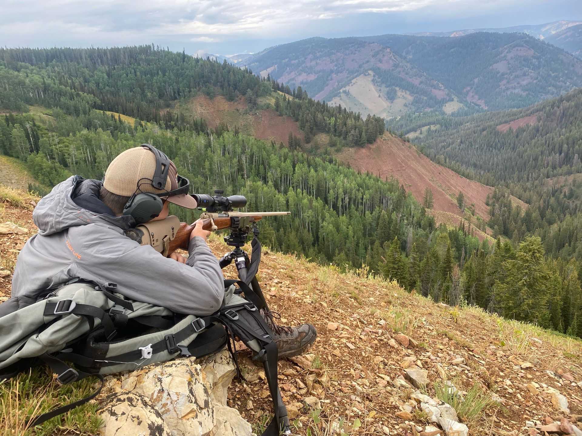 Hunter aiming rifle from a mountain vantage point overlooking a forested valley.