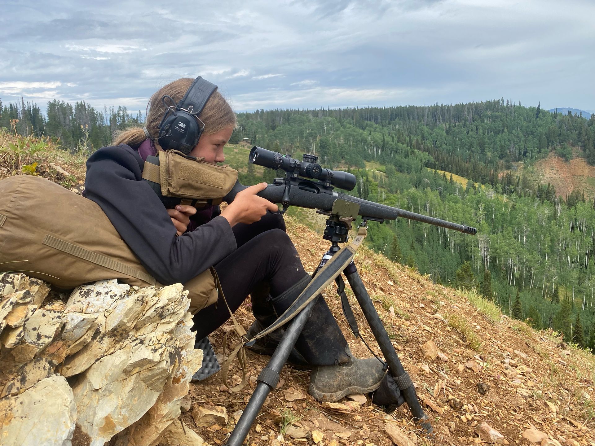 Person aiming a rifle with a scope, wearing ear protection, resting on a hillside with a forested valley.