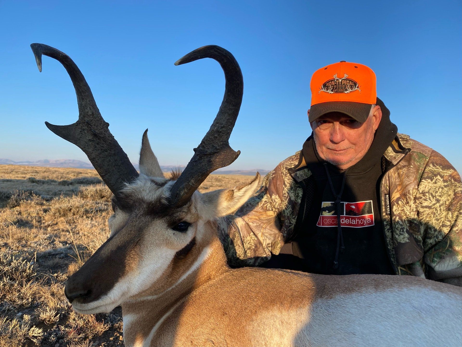 Hunter with a trophy pronghorn antelope, wearing orange hunting hat in a field with a blue sky.