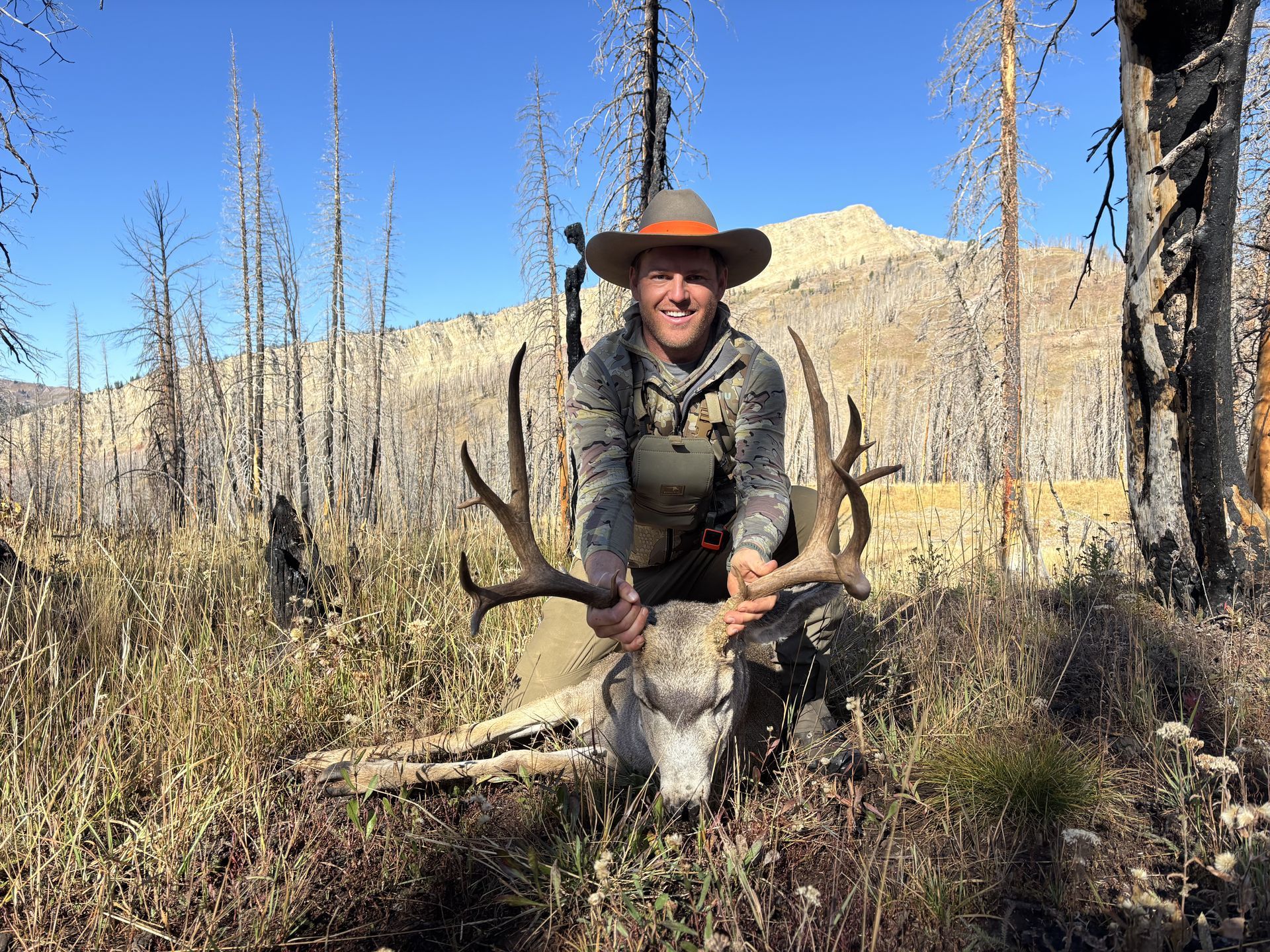 Hunter kneels with large mule deer buck, holding the antlers. Sunlit mountain backdrop, dry grass, burnt trees.