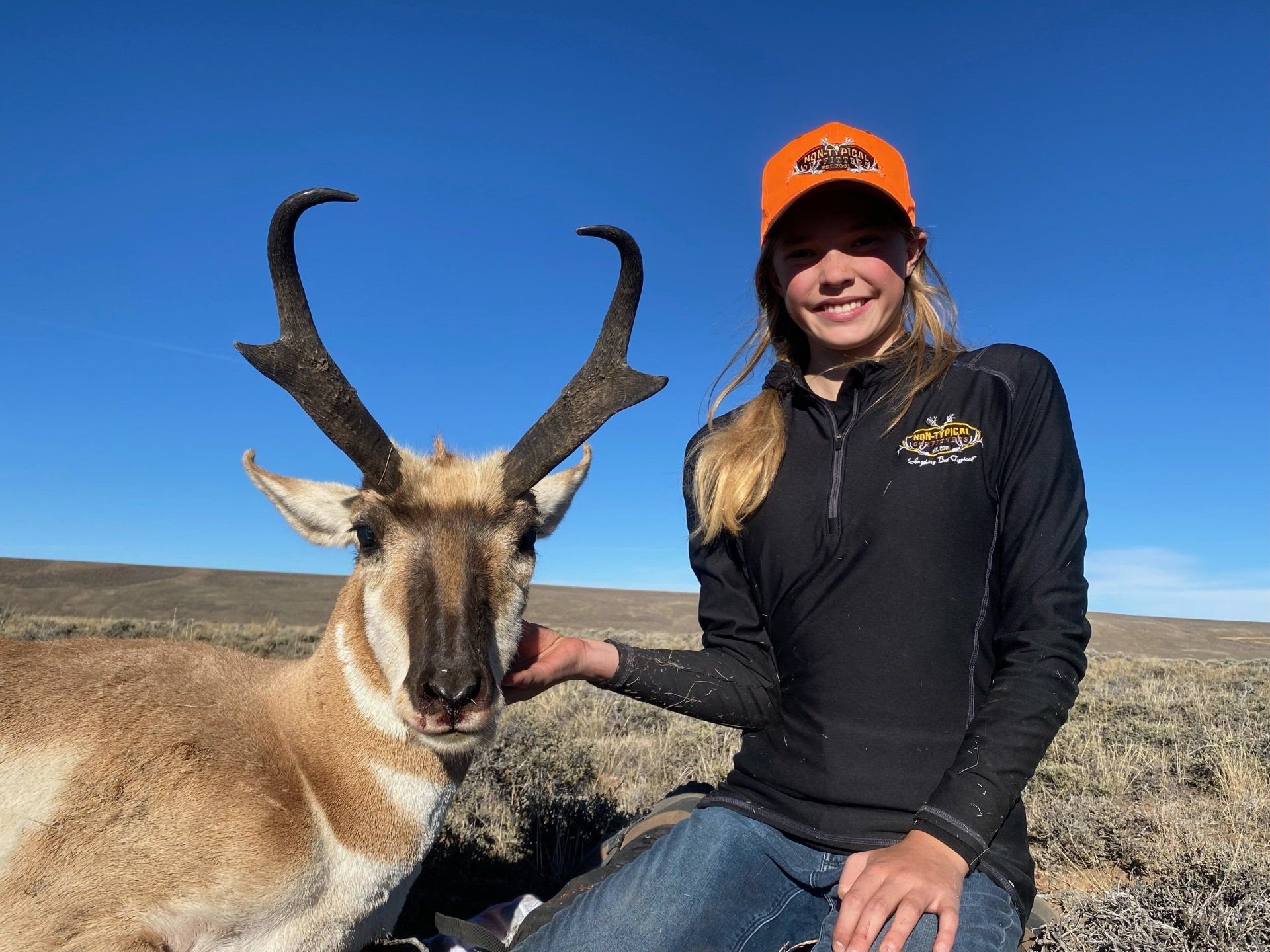 Woman kneels next to a pronghorn antelope in a desert setting; both are facing the camera and smiling.