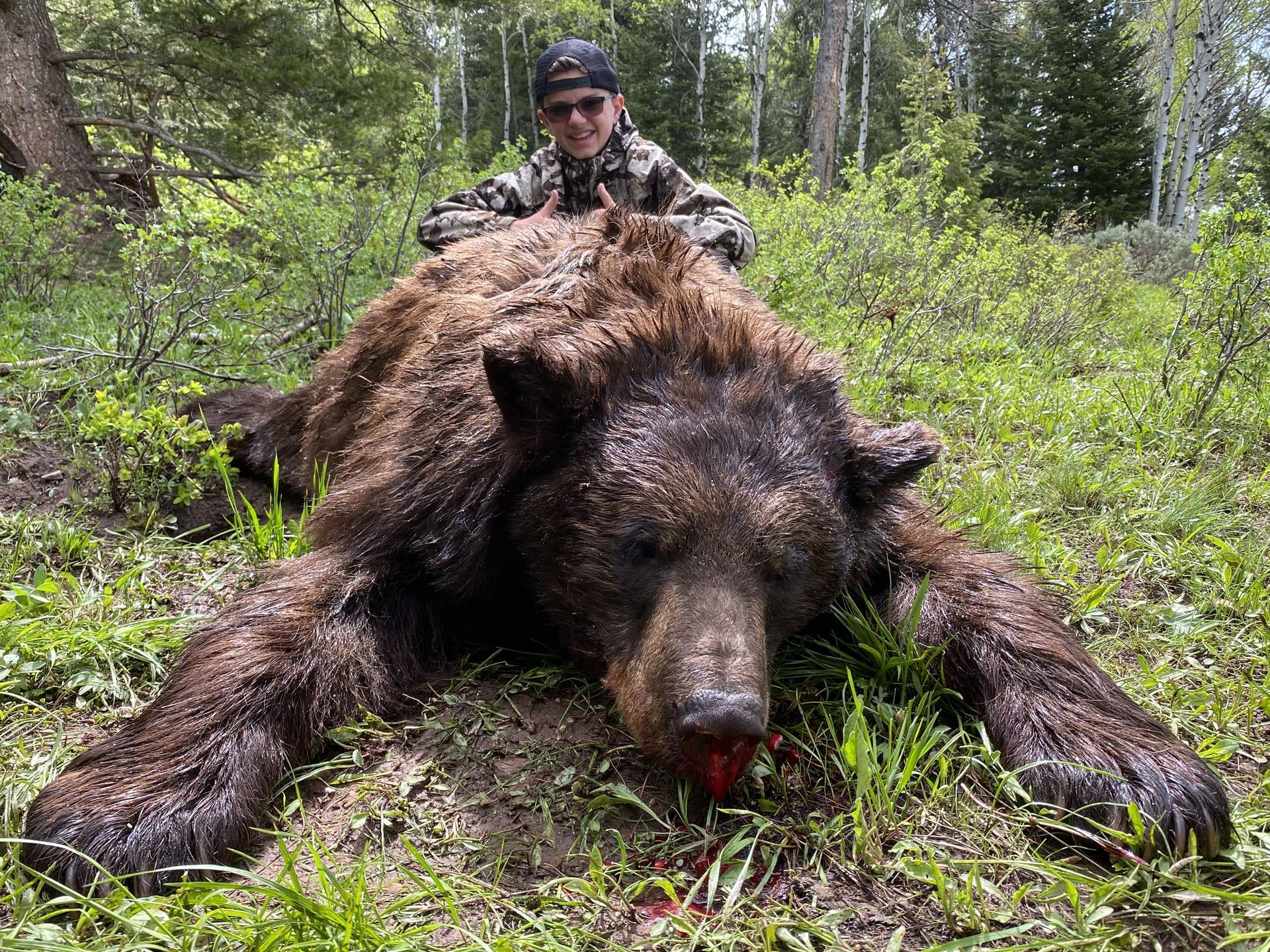 Man in camouflage poses behind a large, dead brown bear lying in grass.