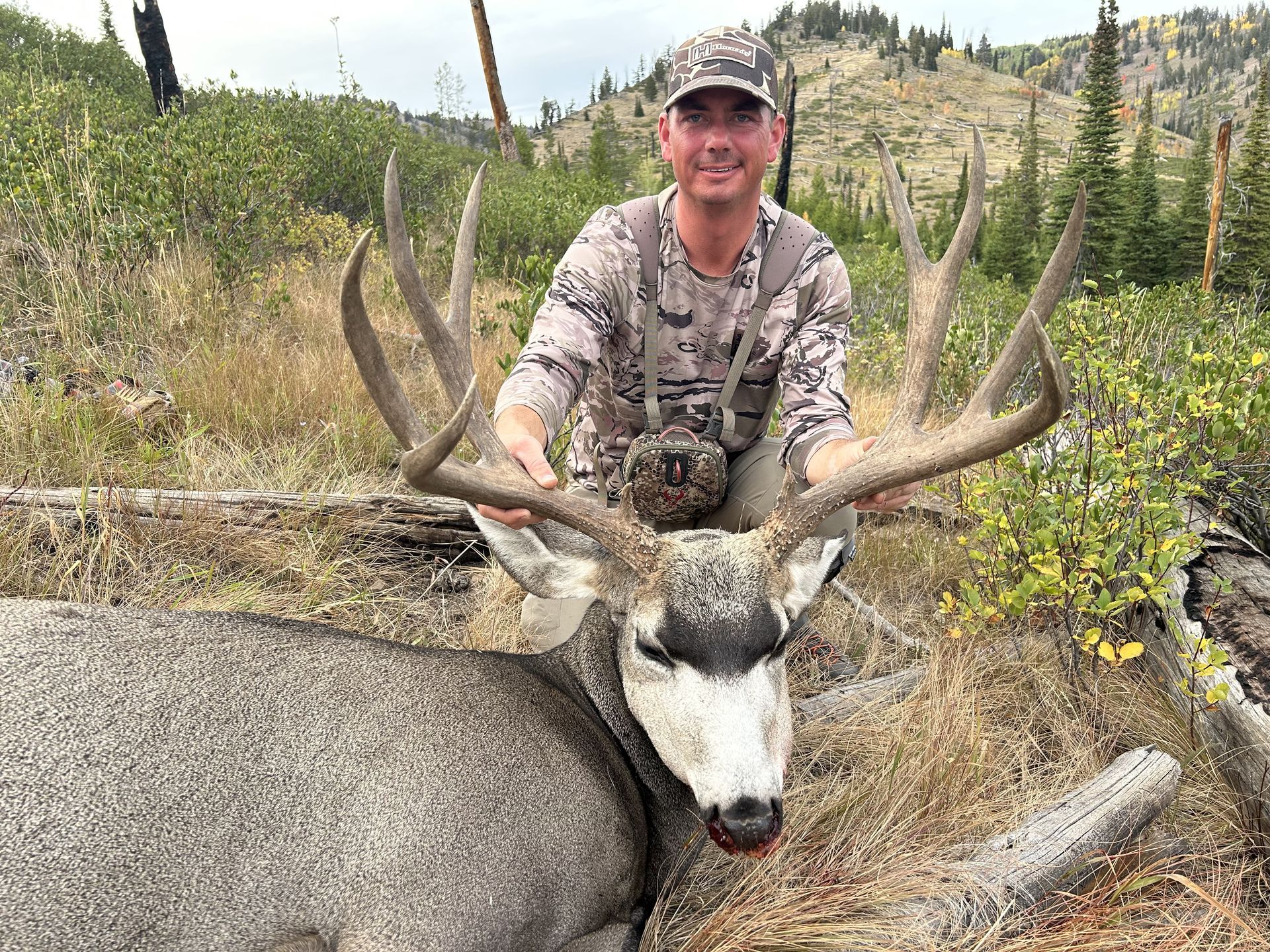 Man in camo holds large buck with impressive antlers in a wooded area.